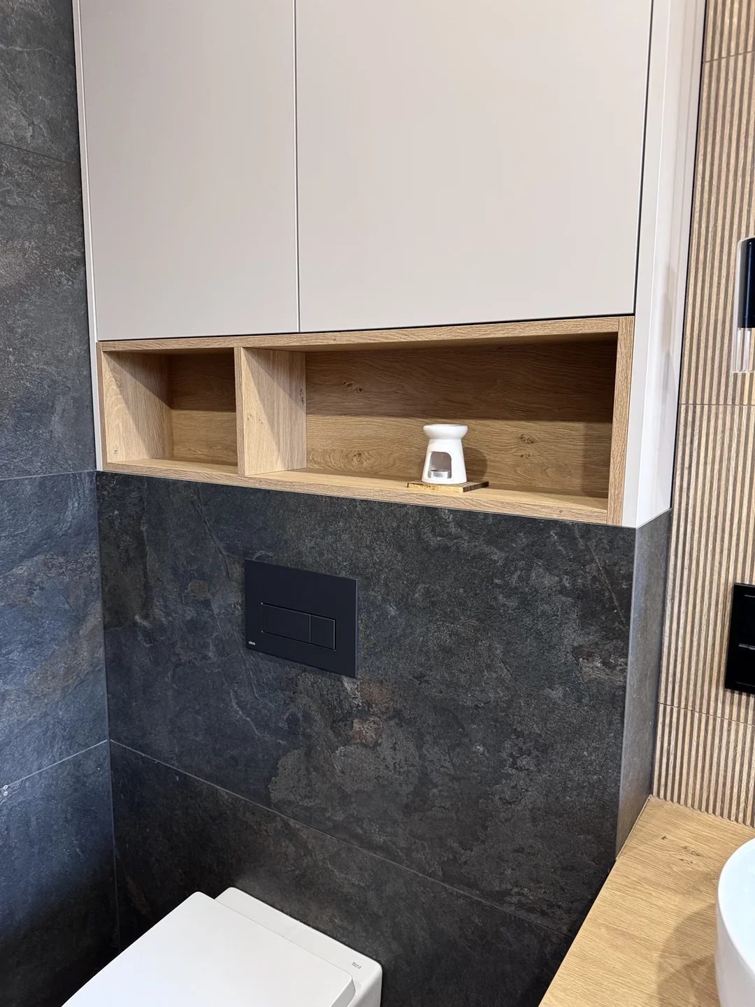 Strefa WC z ciemnymi płytkami, białą zabudową i drewnianą wnęką